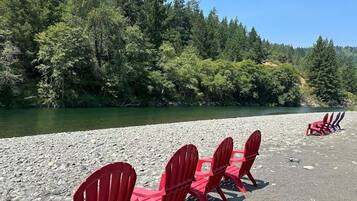 A row of red Adirondack chairs facing a river, surrounded by a rocky beach and dense forest.