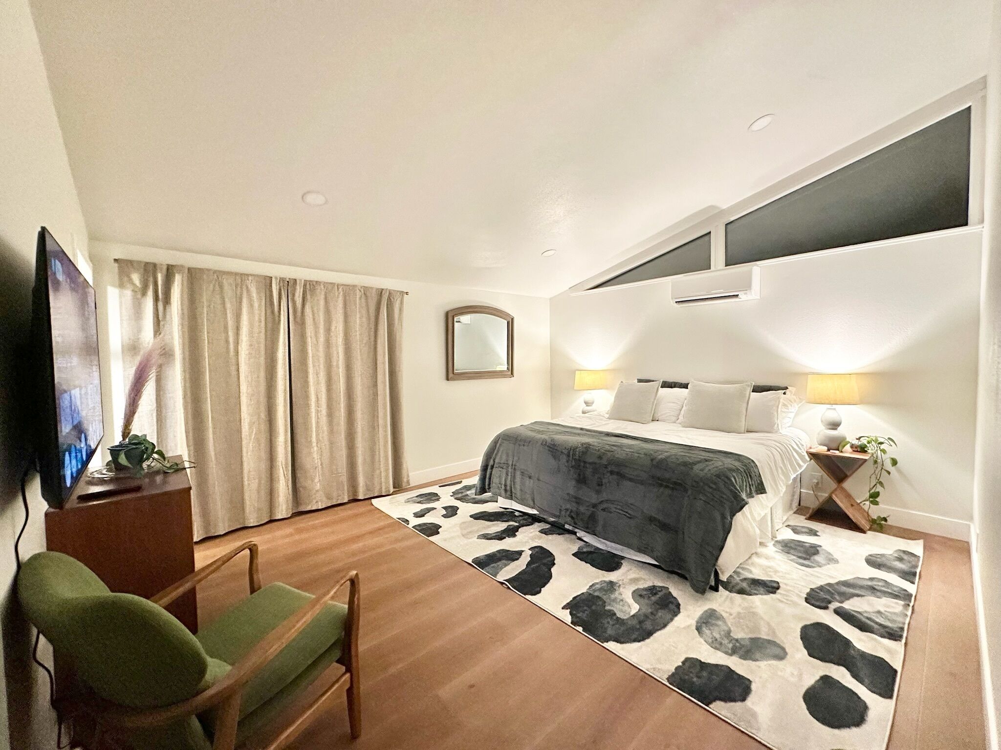 Ett hotellrum med en säng, ett skrivbord med en tv, en stol, ett fönster med gardiner och en spegel.
