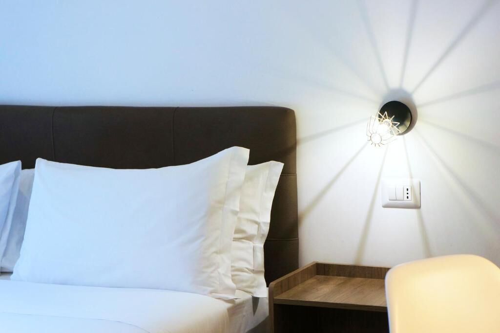En pænt redt seng med hvide sengetøj, en mørk sengegavl og en vægmonteret lampe.