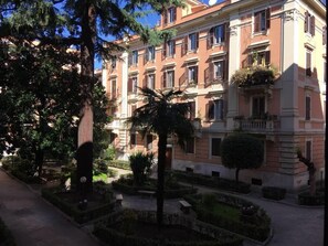 A multi-story building with a symmetrical facade, balconies, and a well-maintained garden with trees and shrubs.