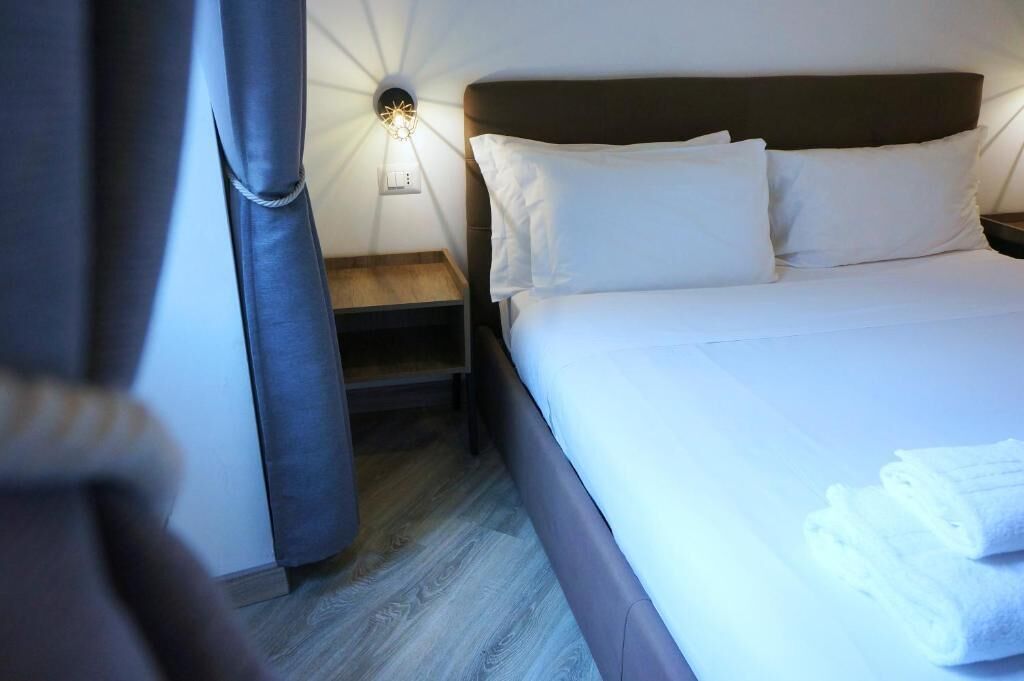 Una cama bien tendida con sábanas y almohadones blancos, una mesita de noche con una lámpara, y una cortina abierta a un lado.