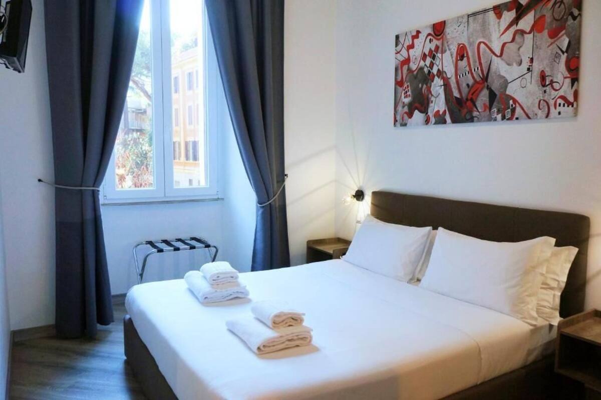Ein Hotelzimmer mit Bett, Handtüchern, Heizkörper, einem Fenster mit Vorhängen und einem Gemälde an der Wand.