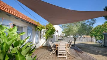Un patio con una mesa y sillas de madera, una casa blanca con techo de tejas rojas y una gran vela de sombra.