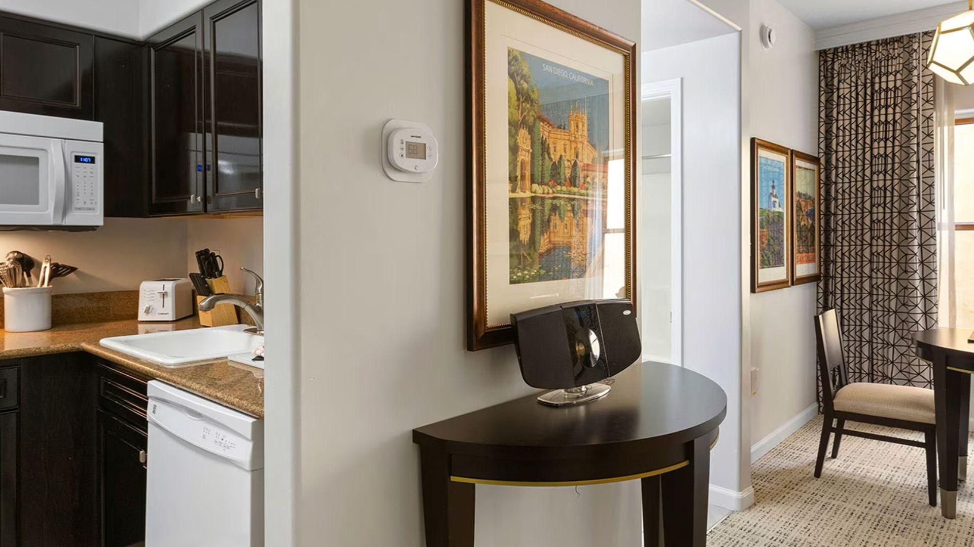A modern kitchen with dark cabinetry, a microwave, a toaster, and a sink. There is a framed picture of a castle on the wall and a small table with a chair.
