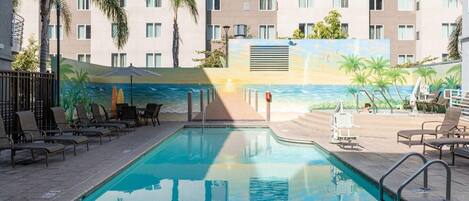 A swimming pool with lounge chairs and umbrellas, surrounded by a building with a mural of a beach scene.