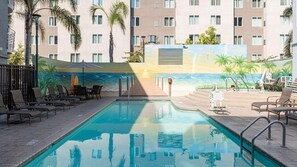 Piscina con sillones de descanso y sombrillas, rodeada de un edificio con un mural de una playa.