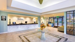 Zona de recepción de hotel con un mostrador de recepción, una mesa con cubierta de vidrio y un arreglo floral, y un área de descanso con vistas al exterior.