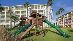 A playground with slides and climbing structures in front of a hotel with multiple balconies and palm trees. - Orlando 2BR Deluxe at Kingstown Reef – Pools, Near Disney & Universal (Orlando)