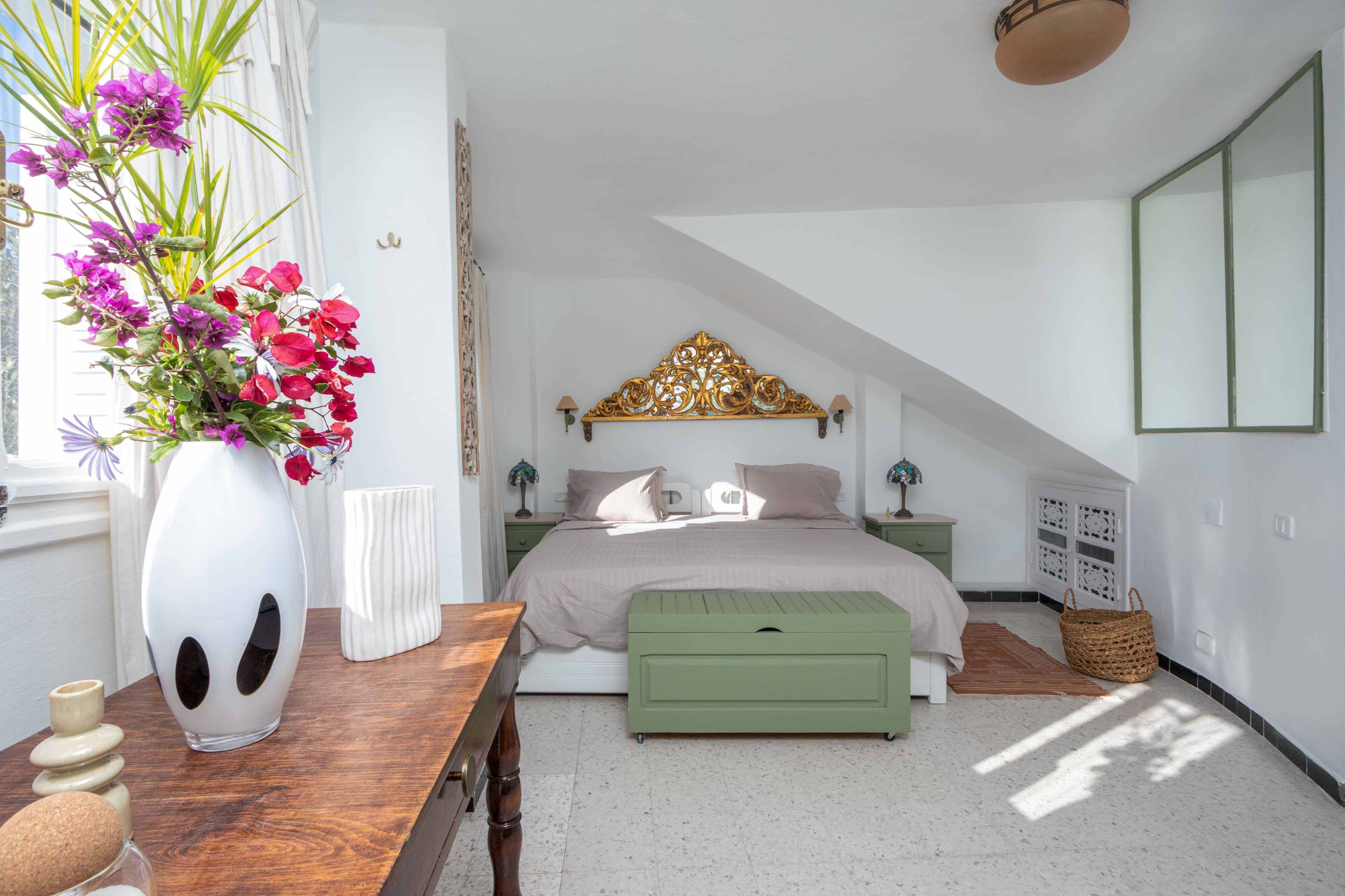 A bedroom with a bed, bedside tables, a dresser, a green bench, a vase with flowers, and a window with curtains.