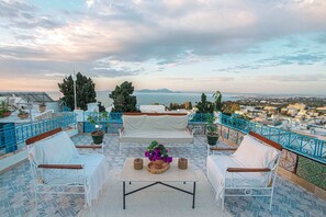 Una terraza con un sofá blanco, dos sillas, una mesa con una planta en maceta y vistas al mar y al paisaje urbano.