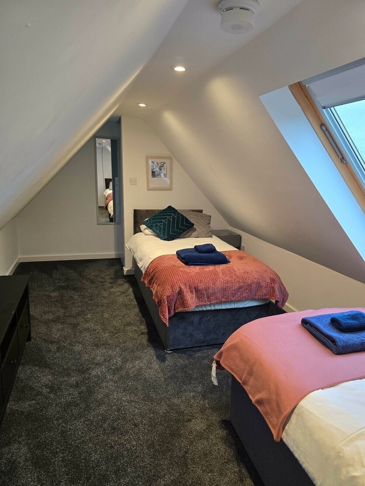 Une chambre avec un plafond en pente, un lit avec une tête de lit, une table de chevet, une commode et une fenêtre.