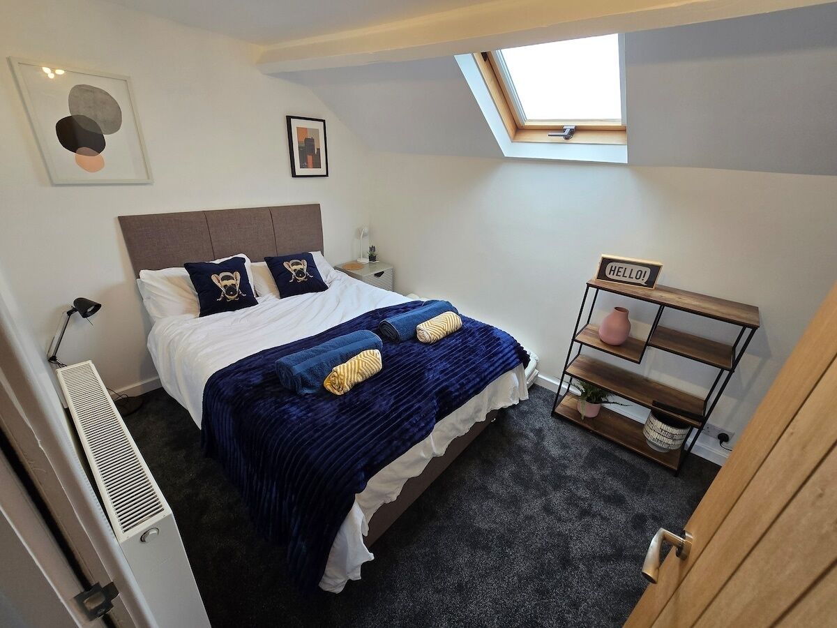A bedroom with a bed, a skylight, a bedside table, a heater, and a wooden shelf with a 'HELLO' sign.