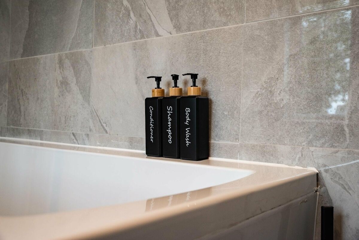 A bathroom with a marble wall and a bathtub. There are three black bottles with pumps on the edge of the bathtub.