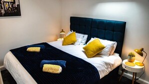 Una cama bien hecha con cabecera azul oscuro, ropa de cama blanca y varios almohadones amarillos. Hay una mesita de noche con una lámpara y una planta en maceta.