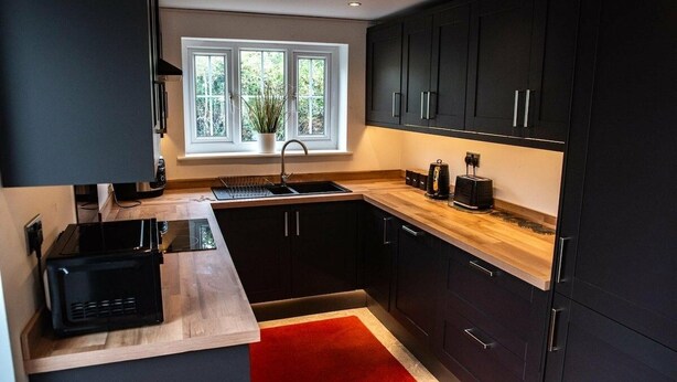 Una cocina moderna con muebles oscuros, encimera de madera, alfombra roja y una ventana con una planta.