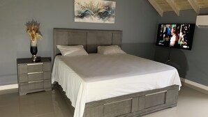 Ein Schlafzimmer mit einem großen Bett, einem Nachttisch mit einer Vase, einem Gemälde an der Wand und einem Fernseher, der eine Fernsehsendung zeigt.