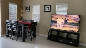 Ein Essbereich mit an der Wand montiertem Fernseher, einem Esstisch mit Stühlen und einer Vase mit Trockenpflanzen.