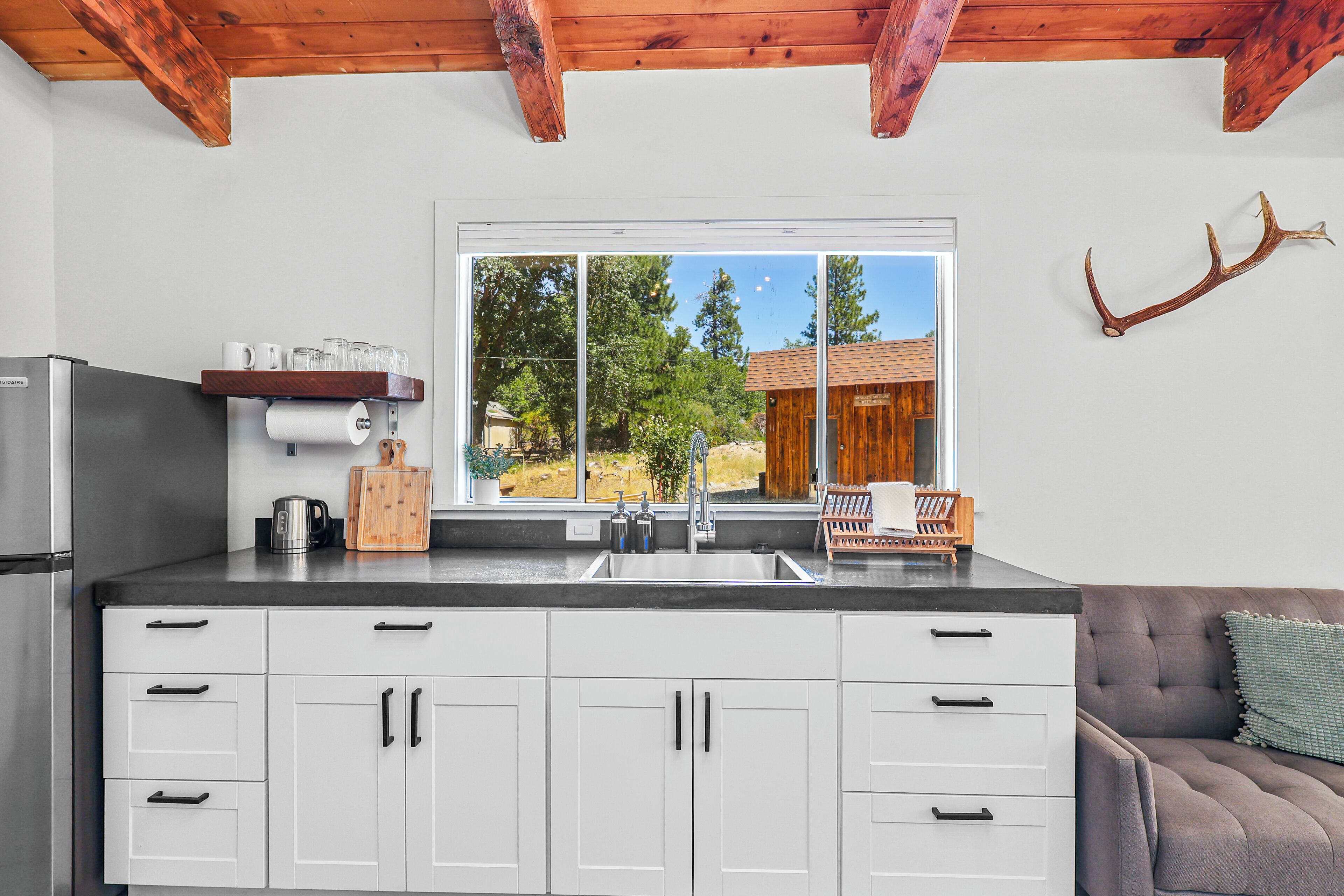 A modern kitchen with white cabinets, a stainless steel refrigerator, and a window overlooking a wooden cabin.