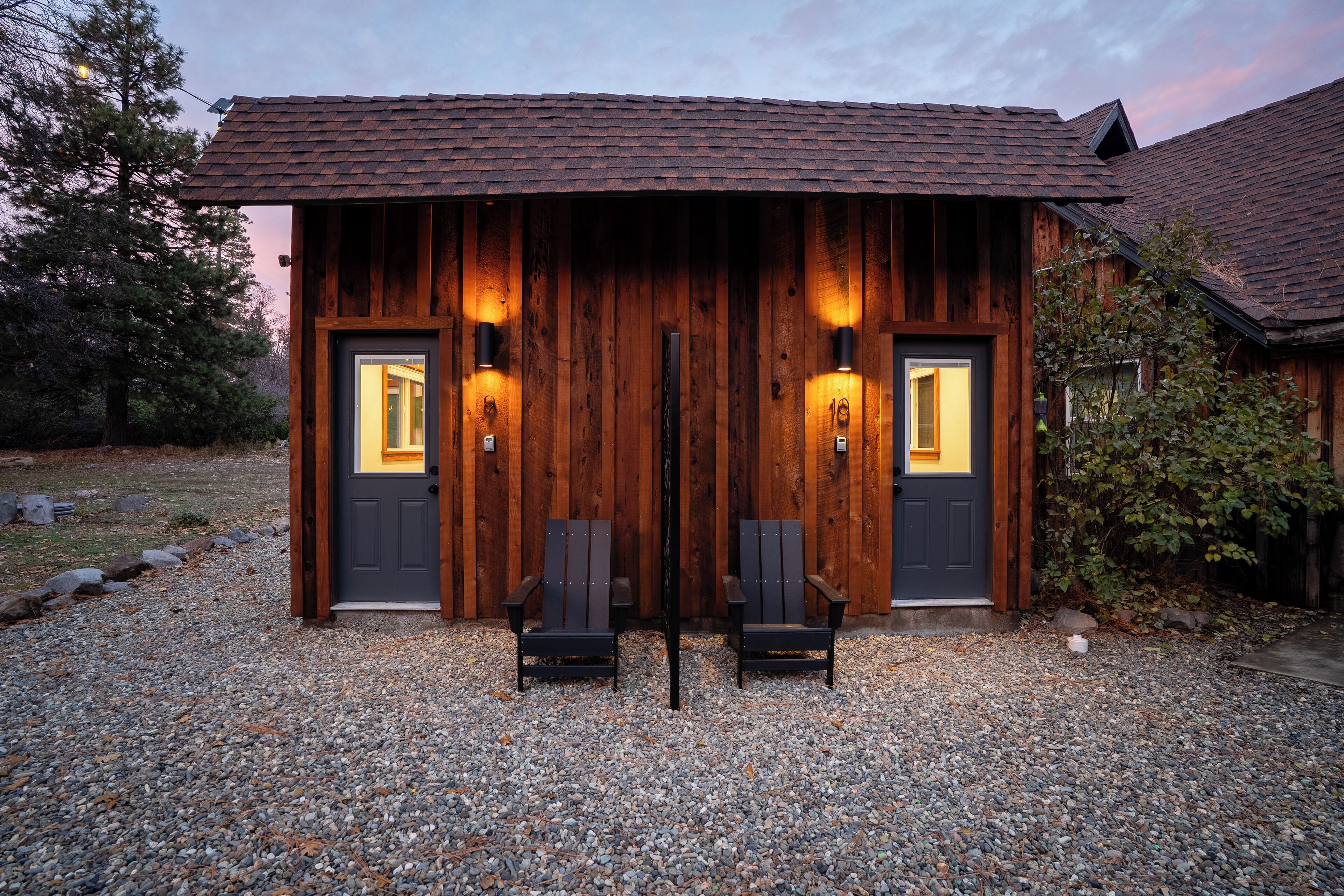 A rustic wooden building with two doors, each with a light fixture. There are two chairs in front of the doors.