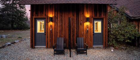 A rustic wooden building with two doors, each with a light fixture. There are two chairs in front of the doors.