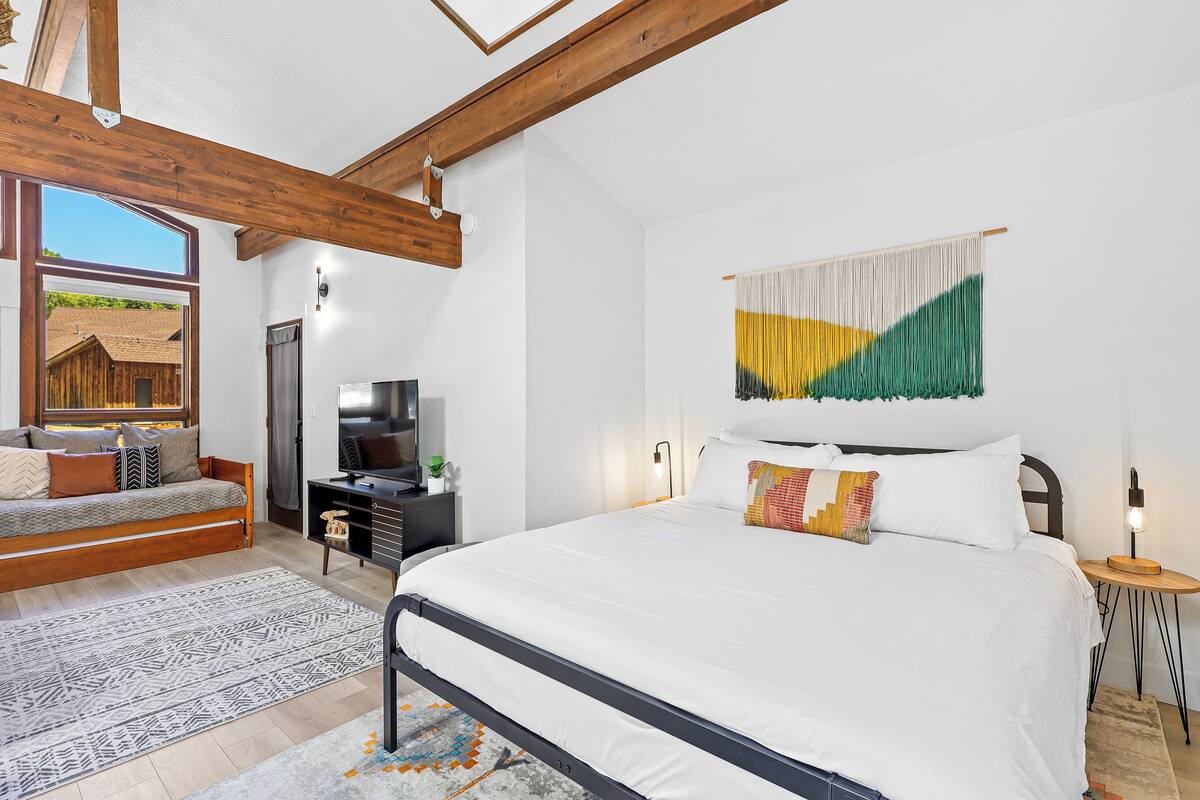 A bedroom with a large bed, a wooden ceiling, a TV, a sofa, and a view of a building outside.