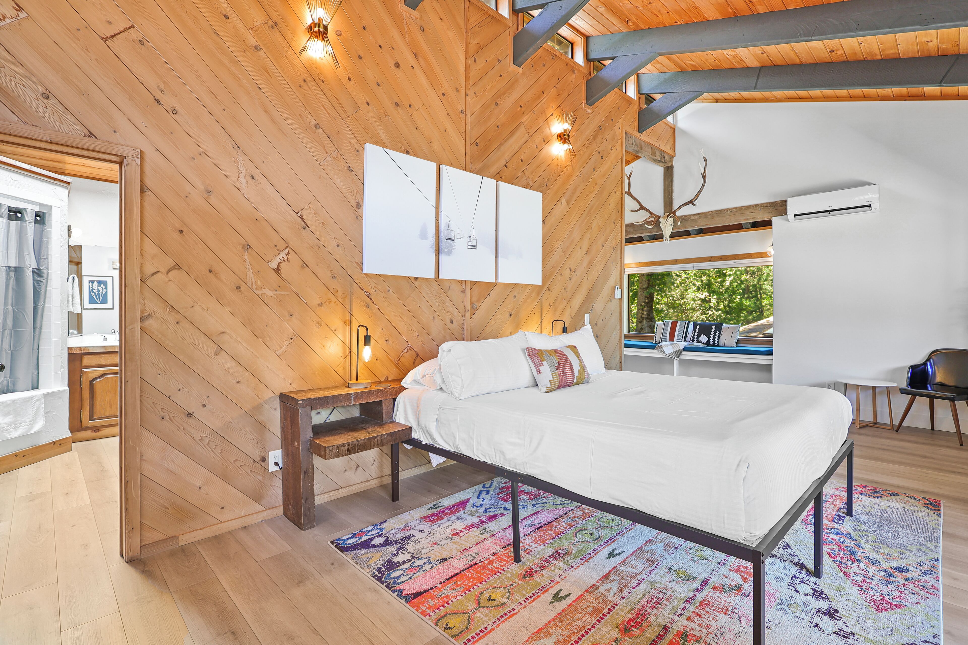 Een slaapkamer met een houten plafond, een bed met wit beddengoed, een kleurrijk vloerkleed en een nachtkastje.