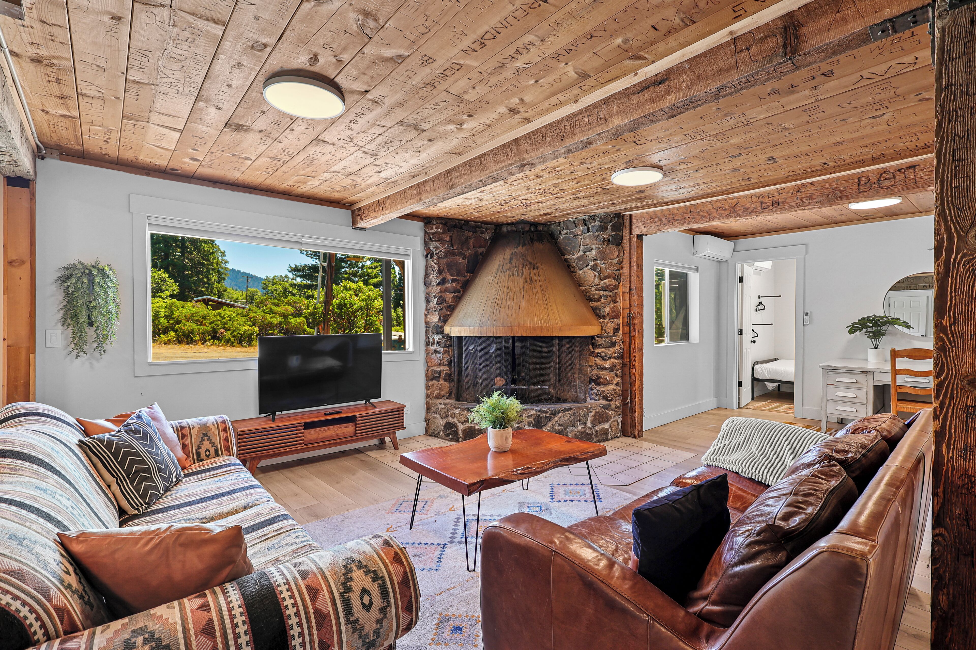 Un salón acogedor con chimenea de piedra, vigas de madera en el techo y un amplio ventanal con vistas a la vegetación.