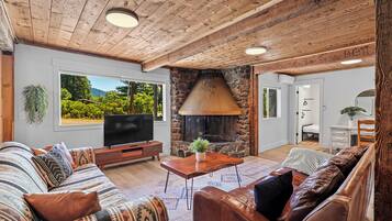 Un salón acogedor con chimenea de piedra, vigas de madera en el techo y un amplio ventanal con vistas a la vegetación.