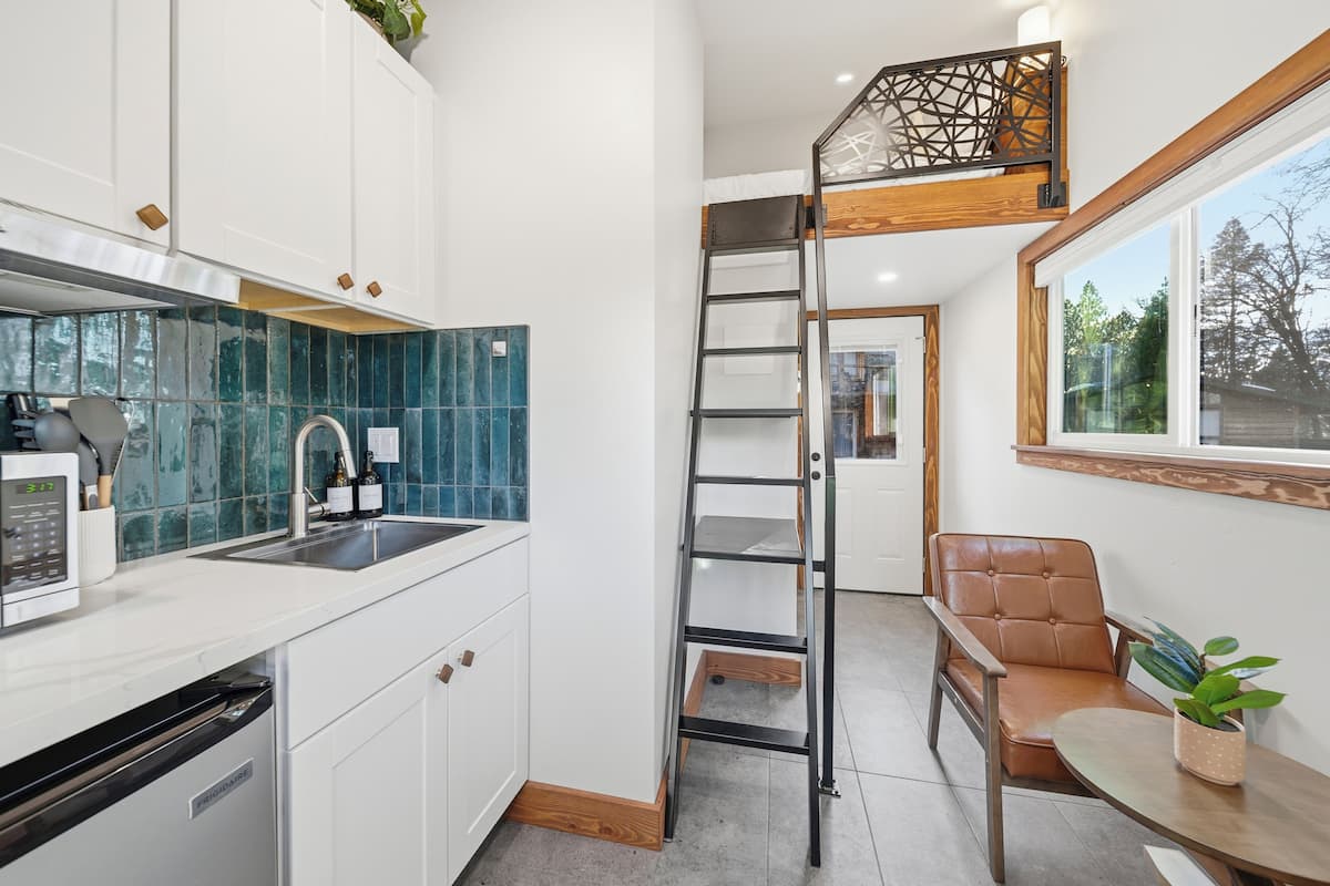 A compact kitchen with white cabinets, a blue tiled backsplash, a stainless steel sink, and a microwave. A wooden ladder leads to a lofted seating area with a brown leather chair and a small table.