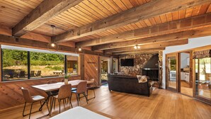 A spacious room with a wooden ceiling, a stone fireplace, a dining area with chairs and a table, and large windows offering a view of the outdoors. - Basecamp Lodge (Mount Shasta)