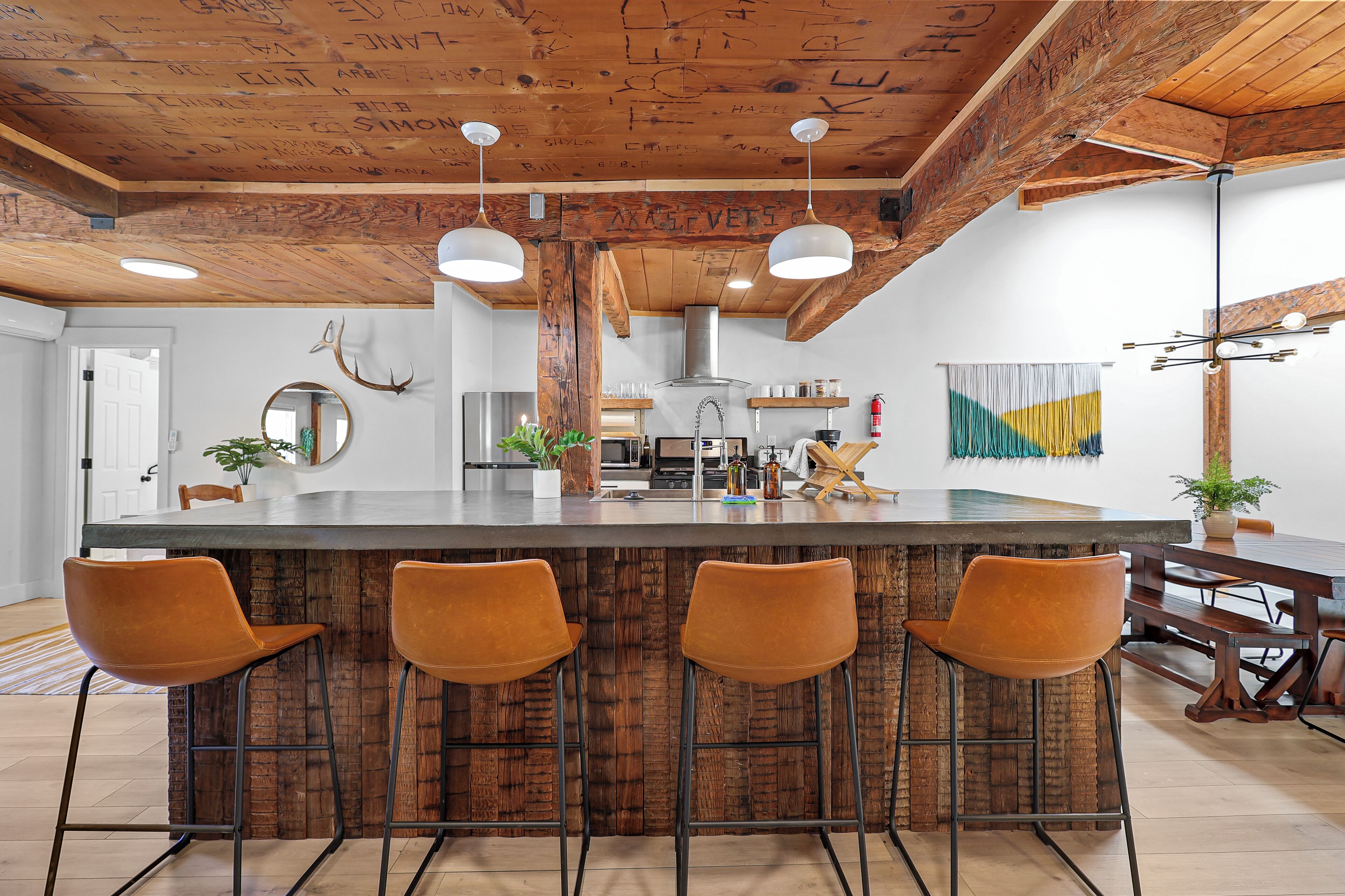 Una cocina moderna con una isla central, taburetes de bar, vigas de madera en el techo e iluminación colgante, además de un colorido tapiz de pared.