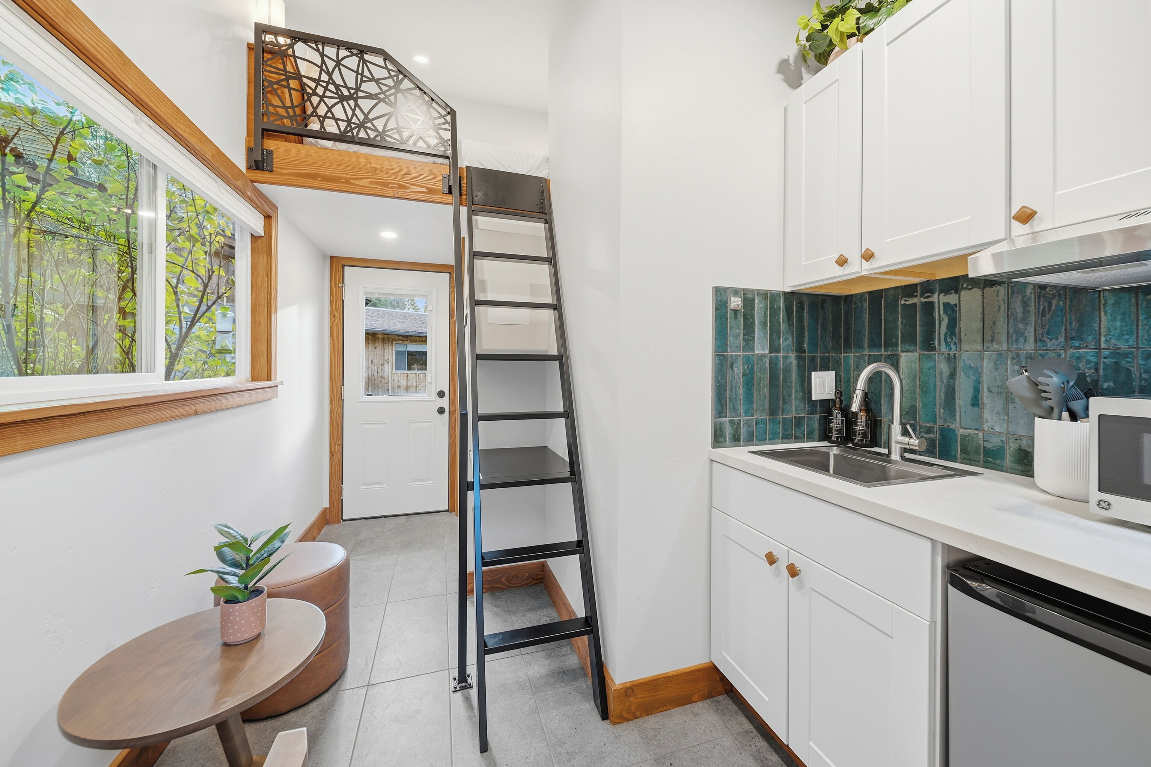 A compact kitchen with white cabinets, a stainless steel sink, and a microwave. A wooden ladder is leaning against the wall, and there's a small round table with a potted plant.