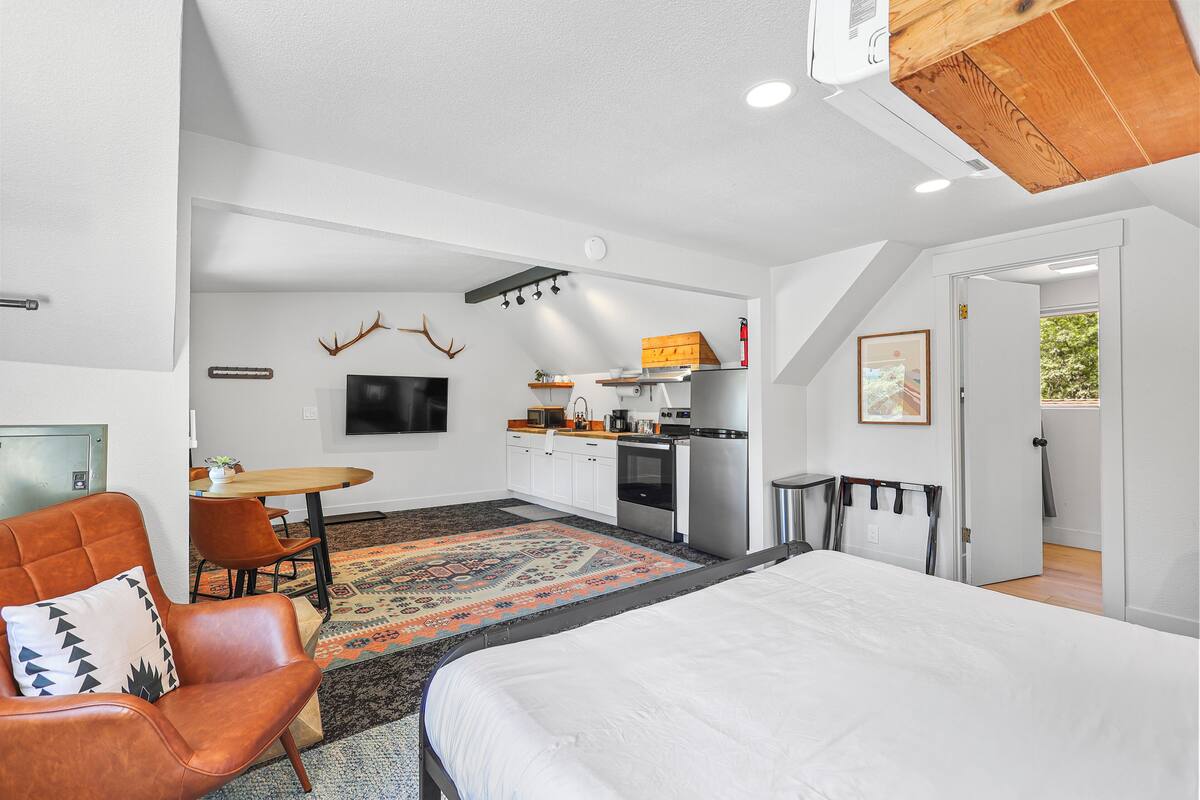 A modern hotel room with a large bed, a dining area, kitchenette, and a view of the outdoors.