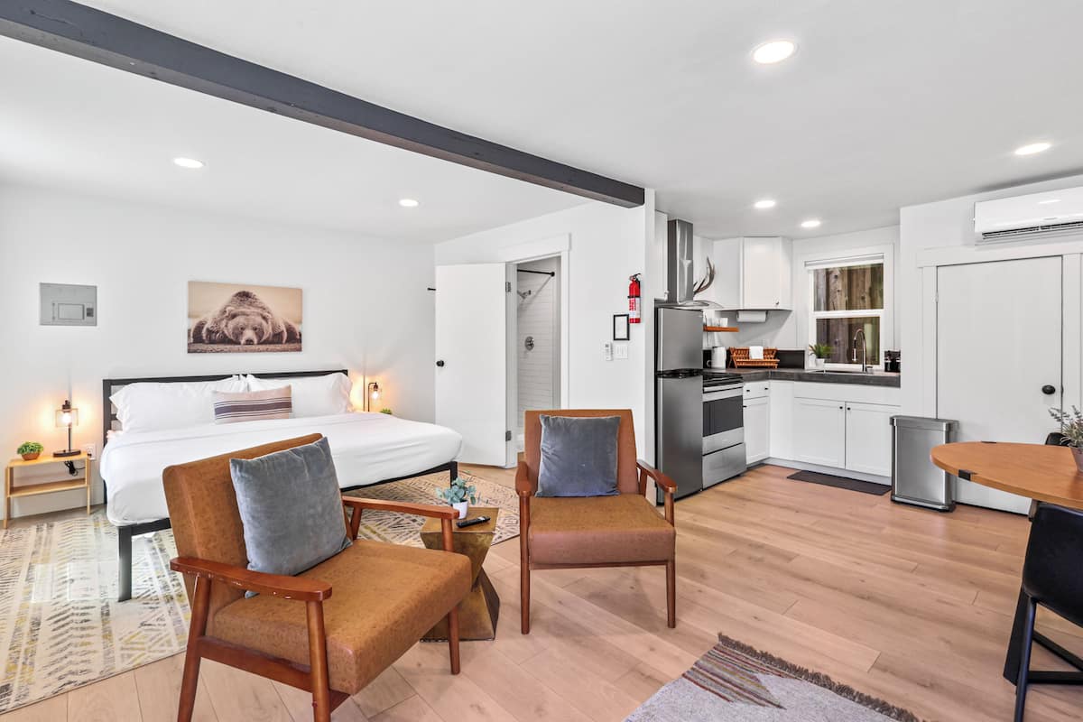 A modern hotel room with a large bed, two armchairs, a small table, and a kitchenette.