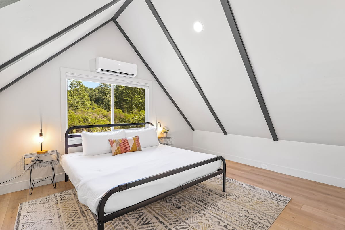 A bedroom with a sloped ceiling, a bed with white bedding, a window with a view of greenery, and a patterned rug.