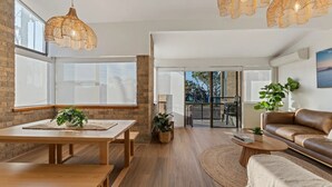 A modern living room with wooden flooring, a brown leather sofa, a dining table with chairs, a hanging light fixture, and a view through the glass door to the outside. - Shoal Bay Beachfront Escape (Shoal Bay)