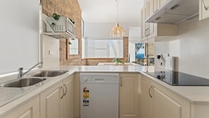 A modern kitchen with a dishwasher, double sink, and a view of the sea through a window. - Shoal Bay Beachfront Escape (Shoal Bay)