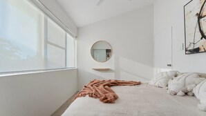 A bedroom with a large window, a round mirror, a bed with white bedding, and a beige throw blanket. - Shoal Bay Beachfront Escape (Shoal Bay)