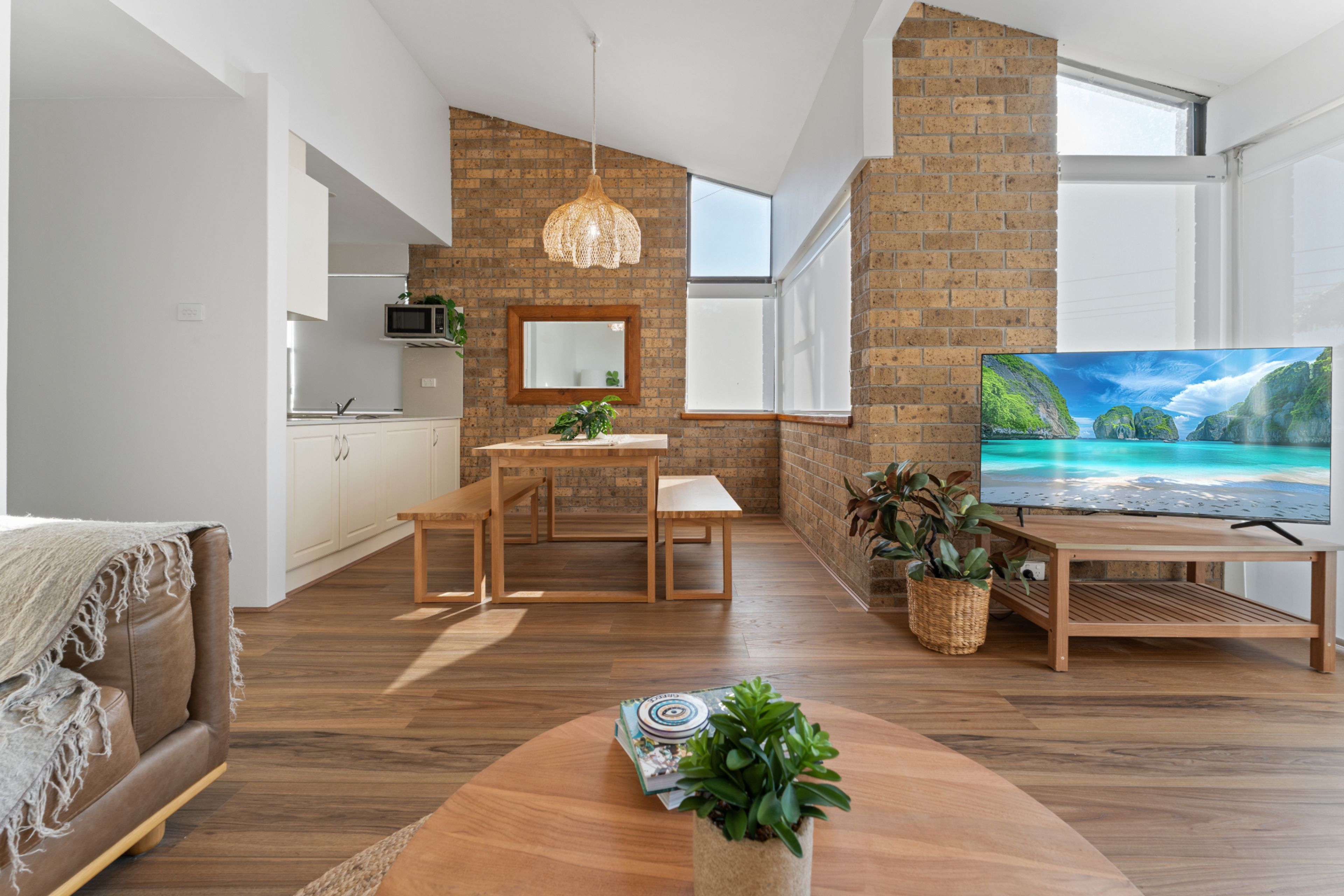 Un salon moderne avec une table basse en bois, un téléviseur posé sur un meuble, un coin repas avec une table et des chaises, une cuisine équipée d’un four à micro-ondes et un mur en briques.