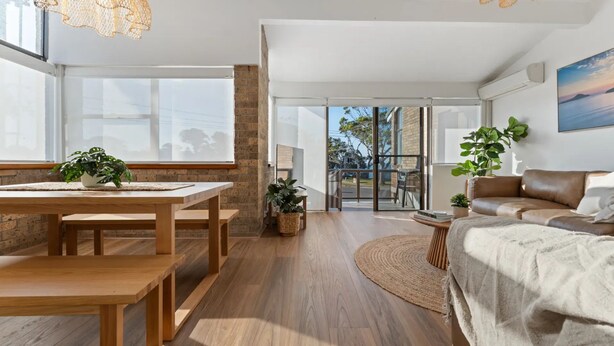 A modern living room with a wooden dining table, a leather sofa, and a large window. - Shoal Bay Beachfront Escape (Shoal Bay)