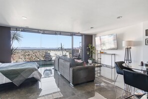 A modern hotel room with a large balcony, a sofa, a dining table, and a TV.