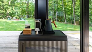 Eine moderne Außenküche mit eingebautem Herd, einer Kaffeemaschine und einem Wasserkocher auf einer Holzterrasse mit Blick auf einen üppigen, grünen Park.
