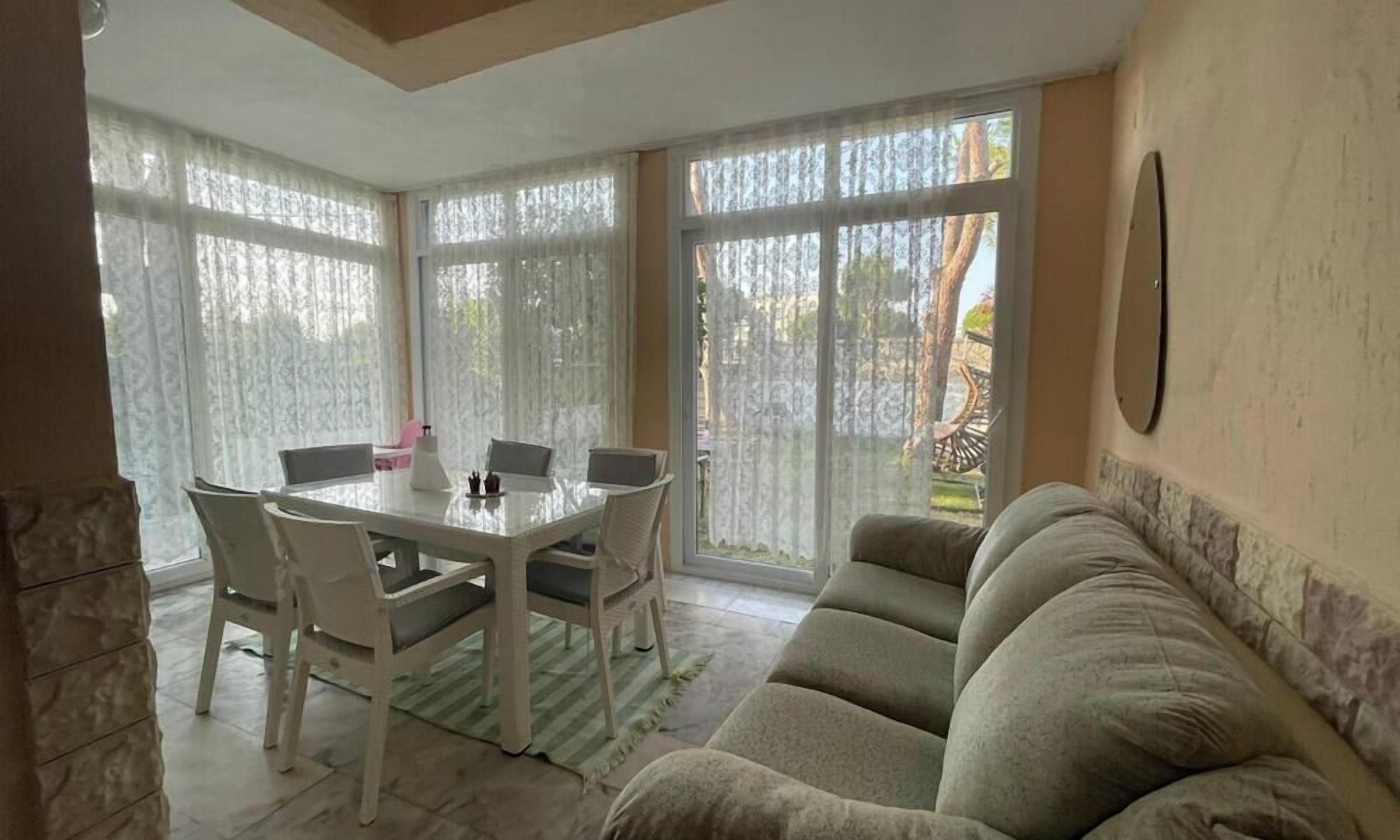 Un comedor con una mesa redonda y sillas, un sofá y ventanas amplias con cortinas traslúcidas.