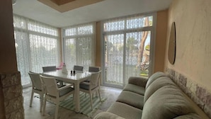 A dining area with a round table and chairs, a sofa, and large windows with sheer curtains.