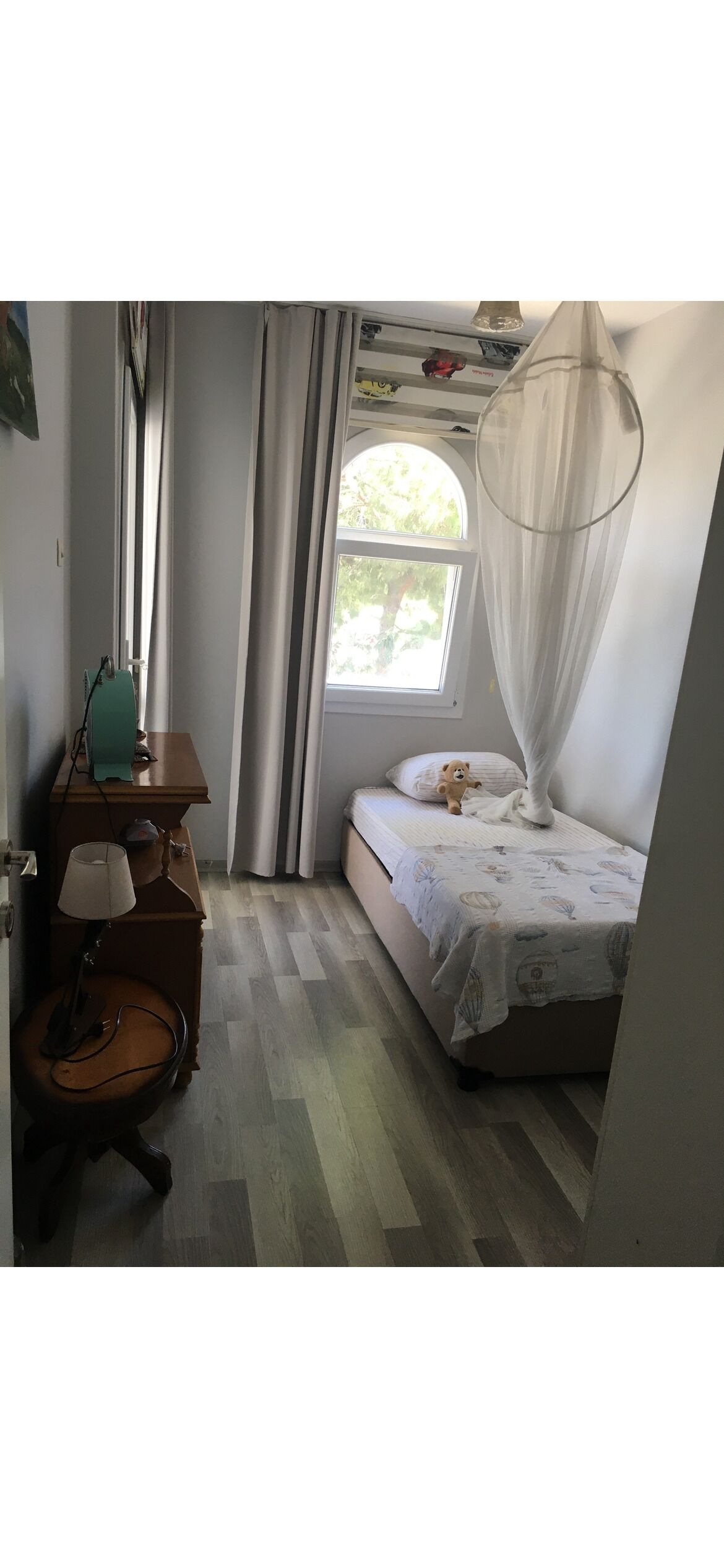 A bedroom with a bed, a bedside table, a lamp, a window with curtains, and a hanging canopy.