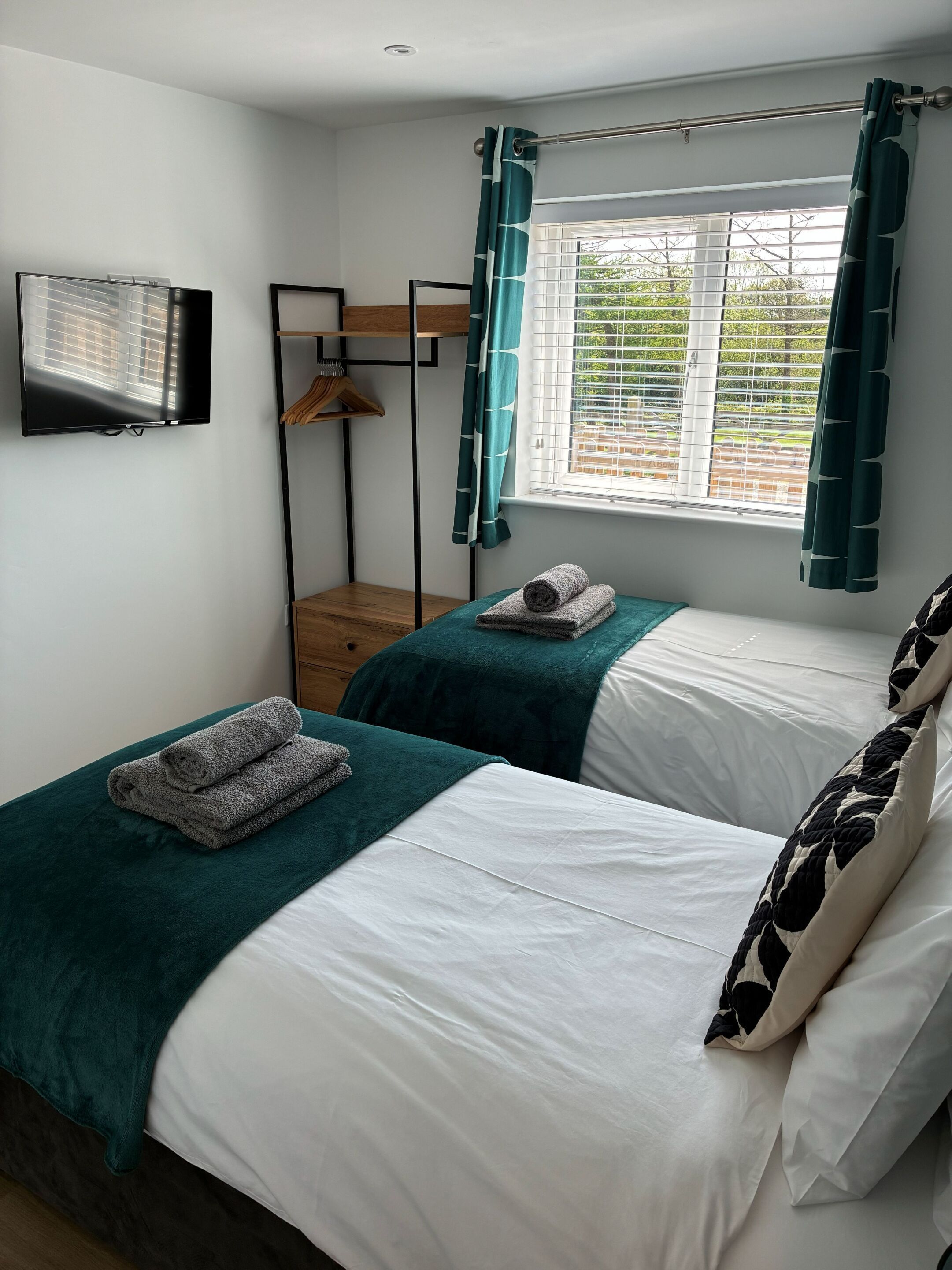 A bedroom with two beds, a TV, a window with curtains, and a wooden shelf.