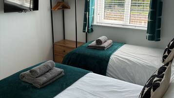 A bedroom with two beds, a TV, a window with curtains, and a wooden shelf.