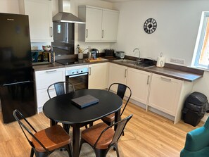 A modern kitchen with white cabinets, a black refrigerator, a stove, and a sink. There is a dining table with chairs and a clock on the wall. - Pet Friendly Cottage with Sea Views (Saundersfoot)