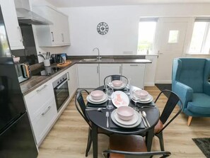 A modern kitchen with white cabinets, a black dining table set with plates, bowls, and cutlery, and a blue armchair in the living area. - Pet Friendly Cottage with Sea Views (Saundersfoot)