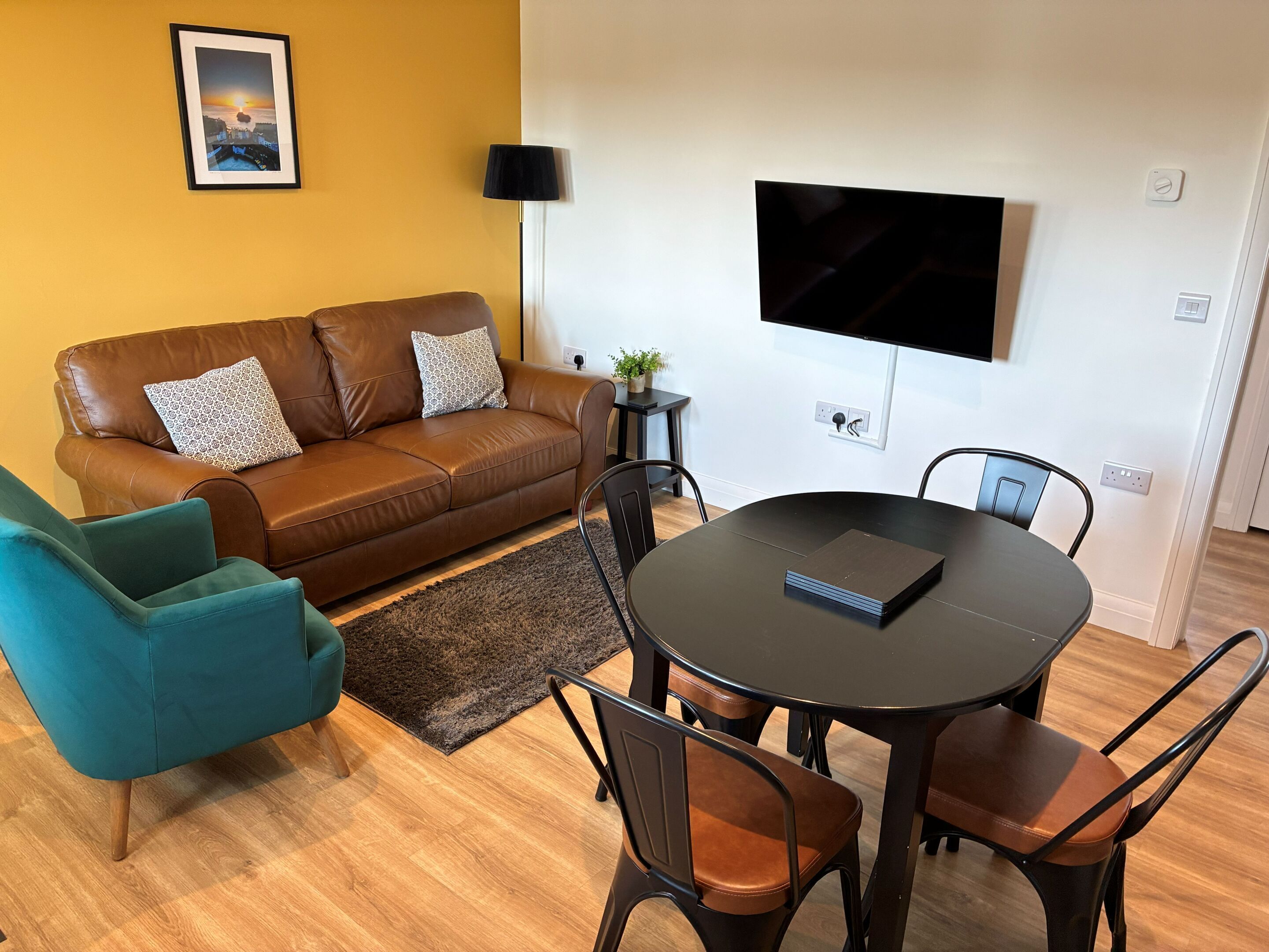 A living room with a brown leather sofa, teal armchair, and a round table with chairs. A flat-screen TV is mounted on the wall.
